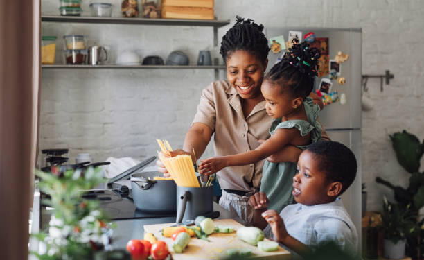 まな板の上でランチのスパゲッティと野菜を準備する母、娘、息子 キッチンのシンクの上でランチのスパゲッティと野菜を準備する幸せなアフリカ系アメリカ人の母、娘、息子。 家族の社会的距離ストック写真や ロイヤリティーフリー画像